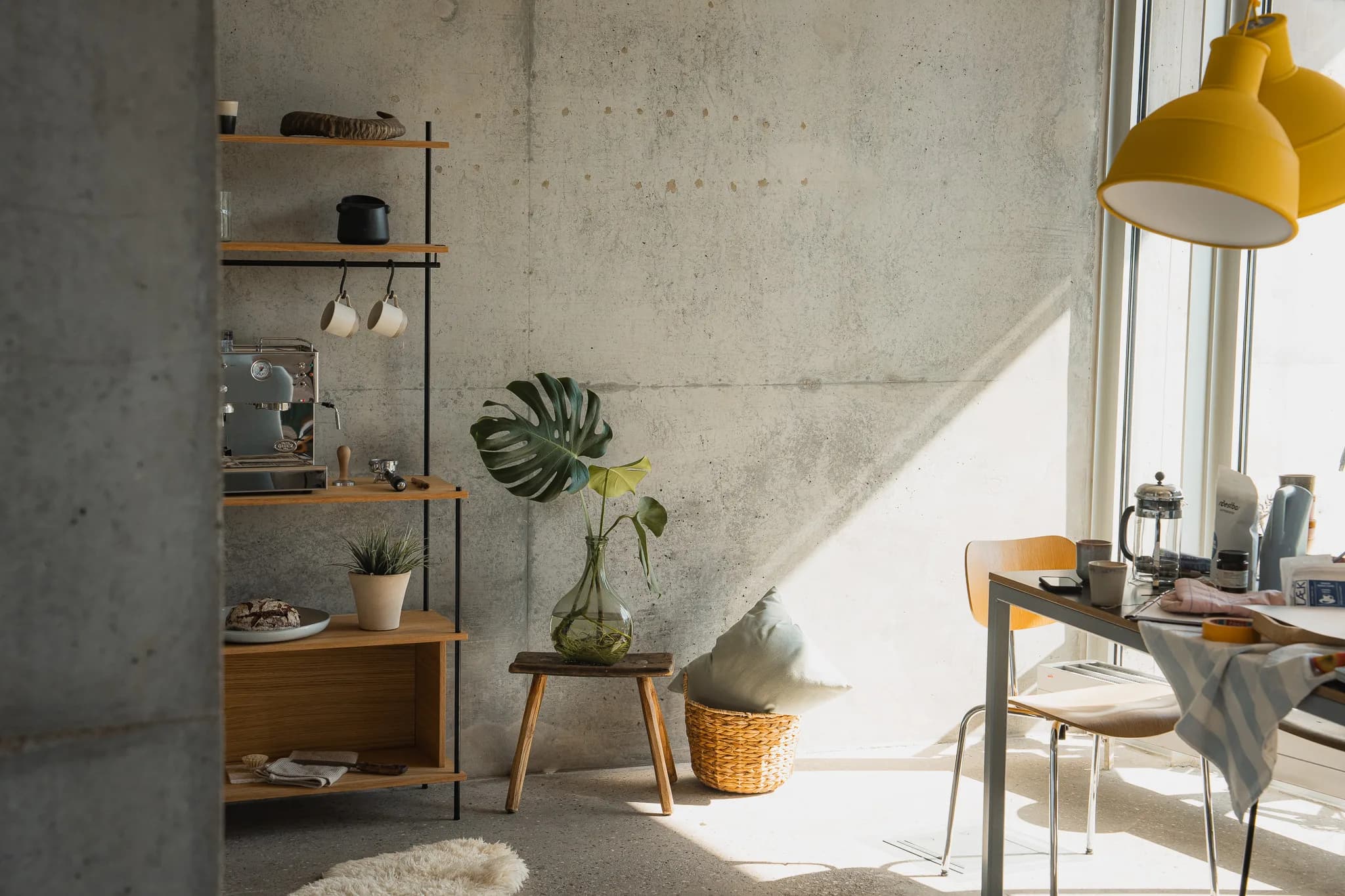 Loft-Küche mit hohem KUUDU-Standing-Regal, links Espresso-Bar, mittig Monstera in Bauchvase und Stuhl-Geflecht, rechts Esstisch im Sonnenlicht